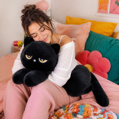 Woman in pink pants and white shirt hugging a black cat plushies on a bed with pink covers and blue and yellow pillows.