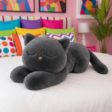 Giant gray cat plushie with closed eyes laying on a bed in a bedroom with colorful pillows, pink walls, and a plant.