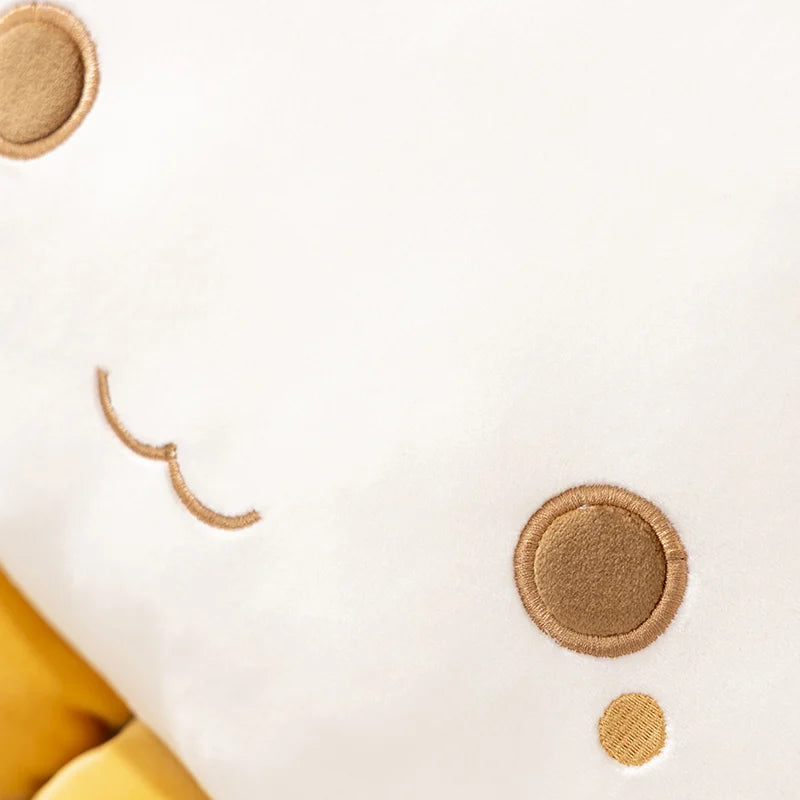 Close-up of a white cat plush with embroidered tan facial features and circular stitched details, including part of a yellow shape at the lower left edge.