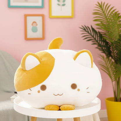 A round, plush cat-shaped cushion or pillow with a white body, yellow patches, and embroidered facial features, displayed on a small white table in a styled room with a pink wall, framed art, and a potted plant.