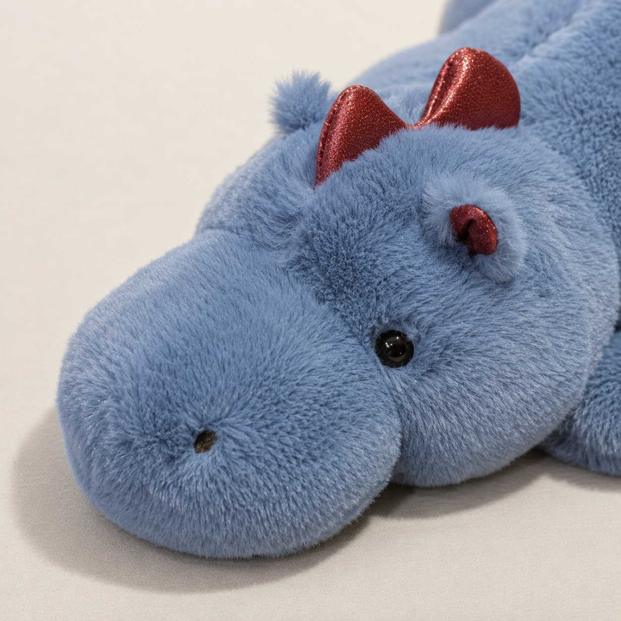 A close-up of a blue plush dragon toy with shiny red ears and small black eyes, lying on a light beige surface.