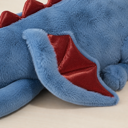 Close-up of a blue plush dragon toy with red spikes and red wing interiors, resting on a light neutral surface.