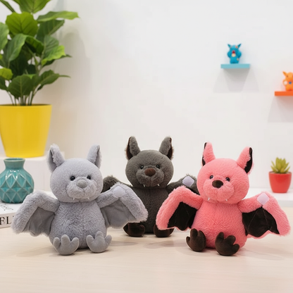 Three plush toys shaped like bats in gray, black, and pink on a light surface with a plant and decorative items in the background.