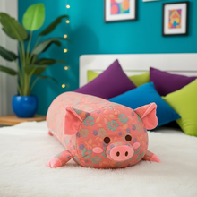 Floral-patterned pink pig-shaped body pillow on a bed with colorful pillows and a plant in the background.