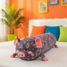 Floral-patterned pig-shaped body pillow on a bed with colorful pillows and an orange wall in the background.