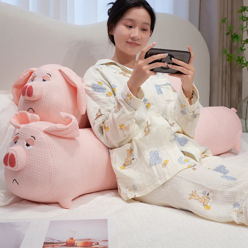Woman in pajamas sitting on a bed with two pink pig-shaped body pillow, holding a phone.