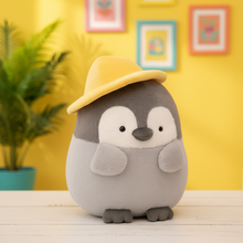 Plush penguin toy wearing a yellow hat on a wooden surface with a yellow wall and plant in the background.