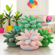 Plush succulent pillows in green and pink on a light wooden floor with colorful plants and a couch in the background.