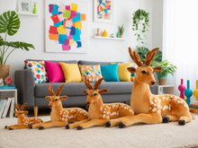 Four plush stuffed deer toys in different sizes arranged on a light beige carpet in a bright, modern living room with a gray sofa and colorful pillows in the background.
