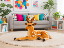 A plush stuffed deer toy with brown fur, white spots, and black hooves is lying on a round beige rug in a bright, modern living room with a gray sofa and colorful pillows in the background.