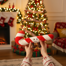 Person holding a candy cane-shaped plush toy in front of a Christmas tree and fireplace.