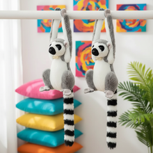 Two plush lemurs hanging from a bar with colorful pillows and abstract art in the background.