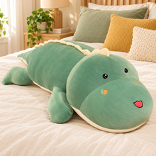 Green oversized dinosaur plush body pillow with open eyes and spiral cheeks on an ivory bed.
