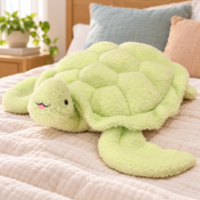 Green Snuggle Shell Turtle Plushie resting on cream bedding with its smiling face and rounded shell visible.